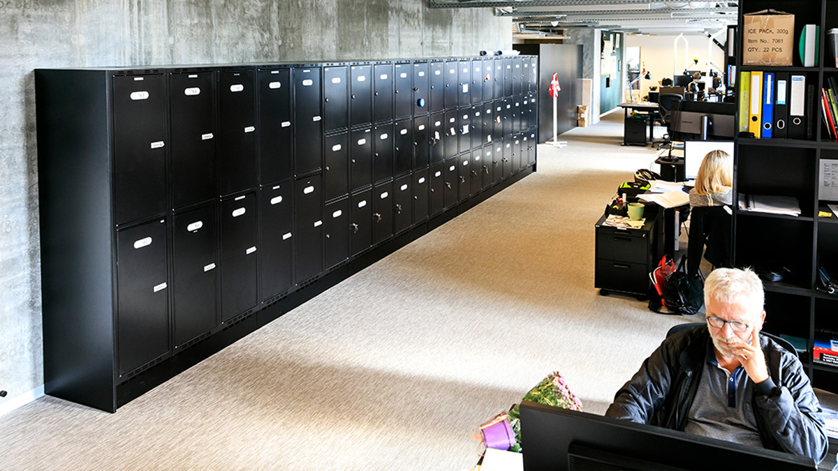 Bilde som vise et todelt garderobeskap plassert inn i et kontormiljø. I forgrunnen ser man en person som sitter og jobber ved en datamaskon.