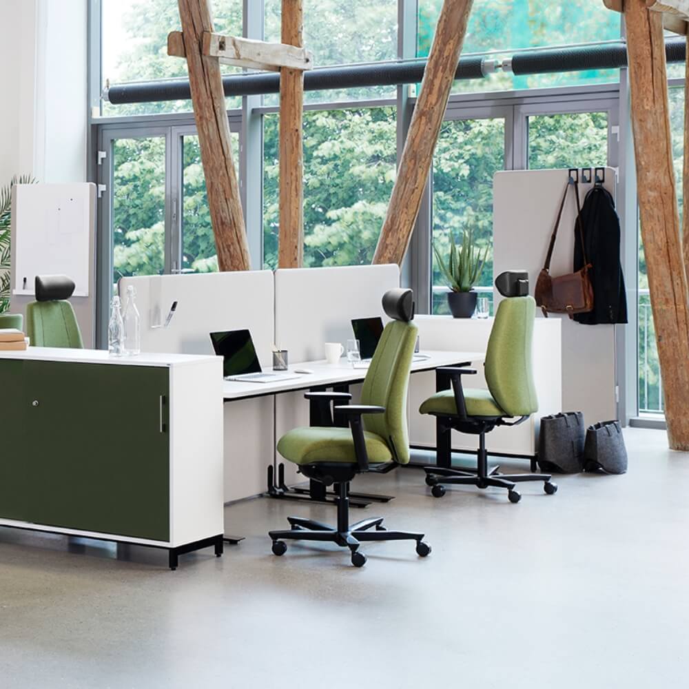 Et åpent kontorlandskap med grønne kontorstoler, hvite bord med skillevegger, store vindu og store trestolper midt i lokalet.
