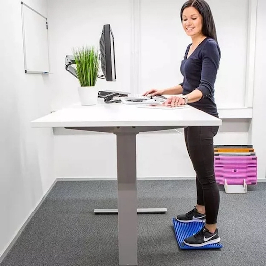 En kvinne står oppreist foran pulten sin og jobber, på en blå avlastningsmatte inne på kontoret.