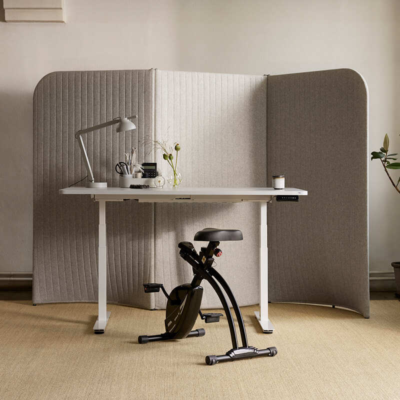 Skrivebordsykkelen BikeIt 2.0. står forann en hvit pult og en grå skillevegg på et lyst hjemmekontor.