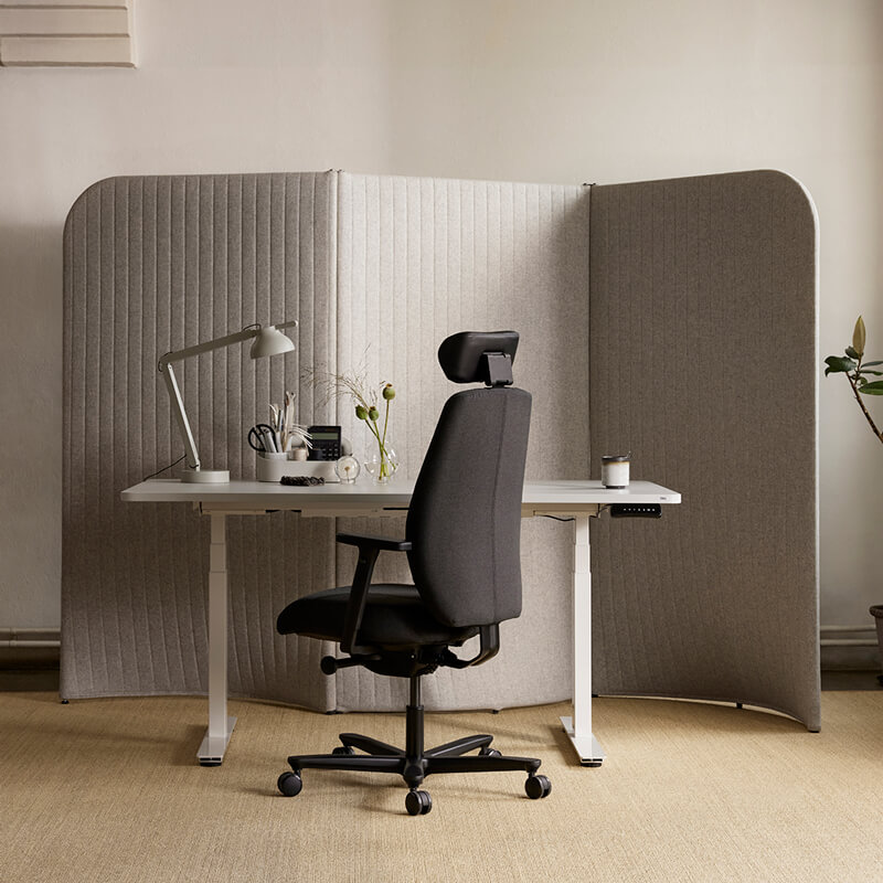 En ergonomisk kontorstol står forann en hvit pult og en grå skillevegg på et lyst hjemmekontor.