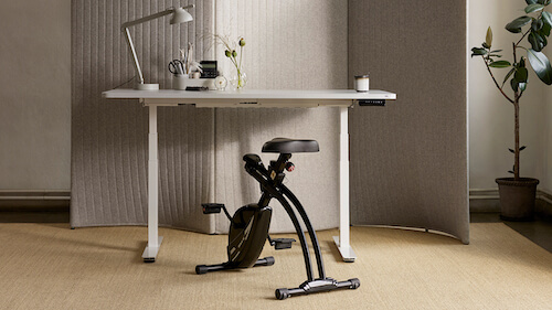 Bike it 2.0 står inntil et hvitt skrivebord med lampe på et hjemmekontor med tregulv og lyse vegger.